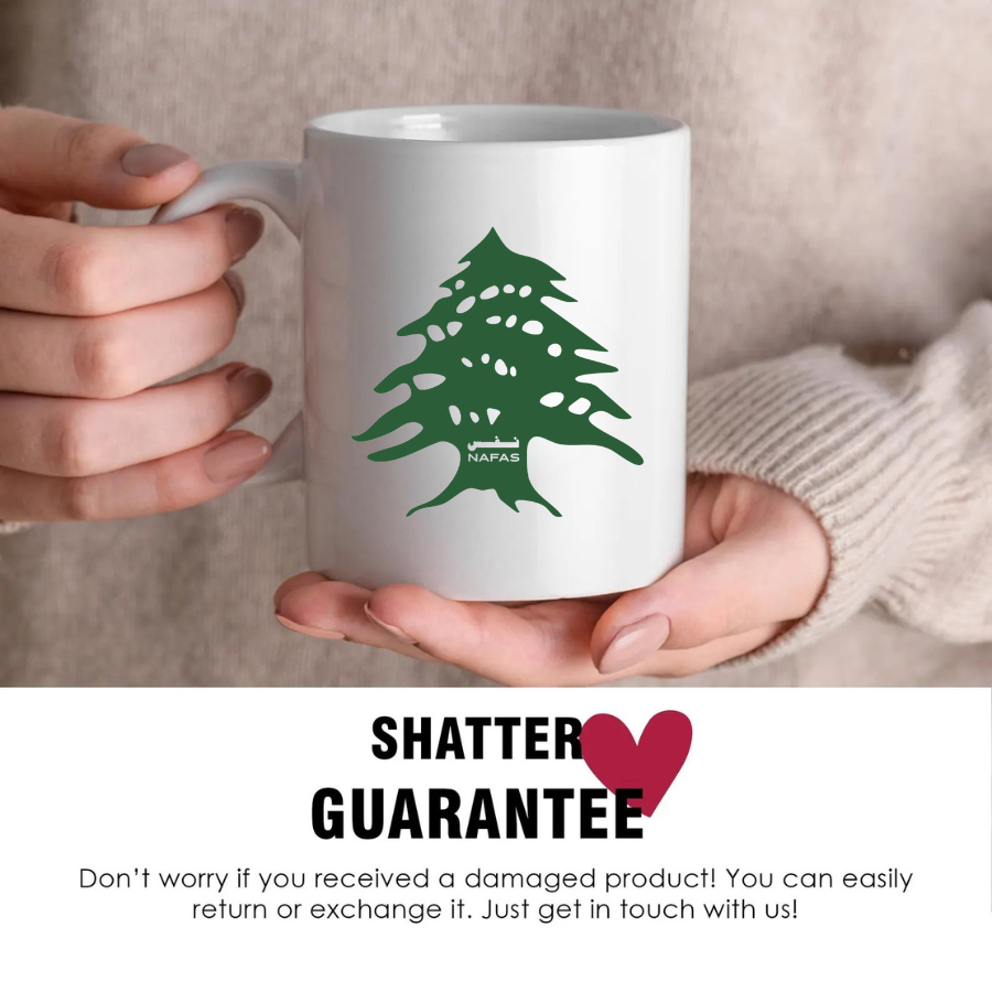 Hands holding a white 11 oz ceramic mug with green Lebanese cedar tree design by Nafas, showing shatter guarantee message
