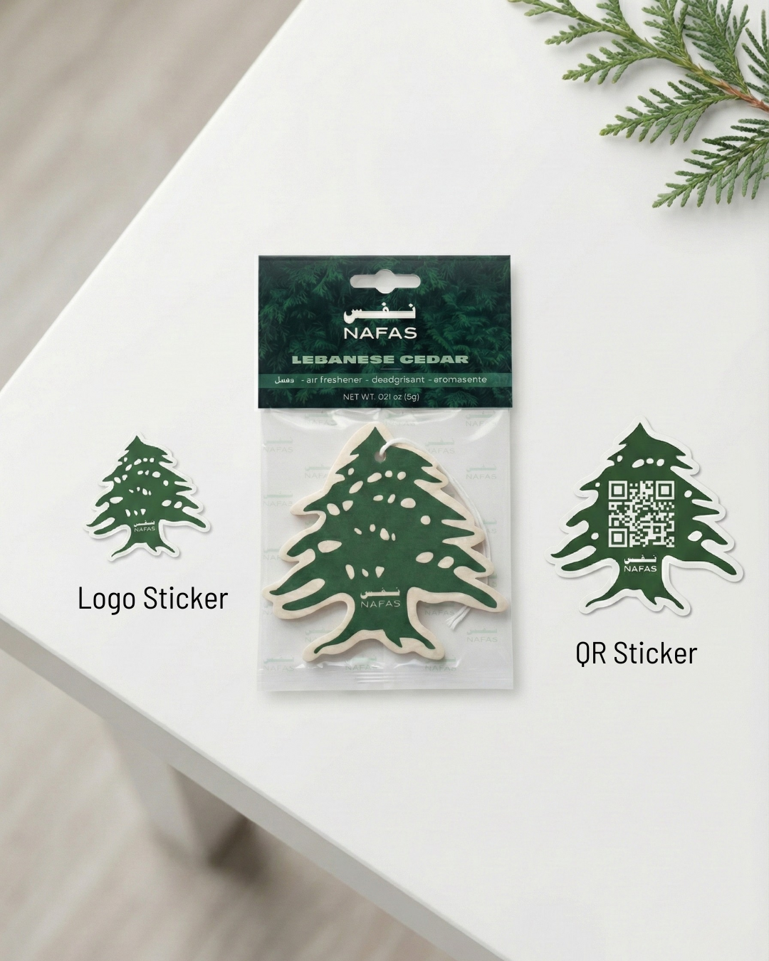 Flat lay of a Nafas Lebanese Cedar car air freshener in its green packaging on a white table, with a free cedar logo sticker on the left and a cedar QR code sticker on the right.