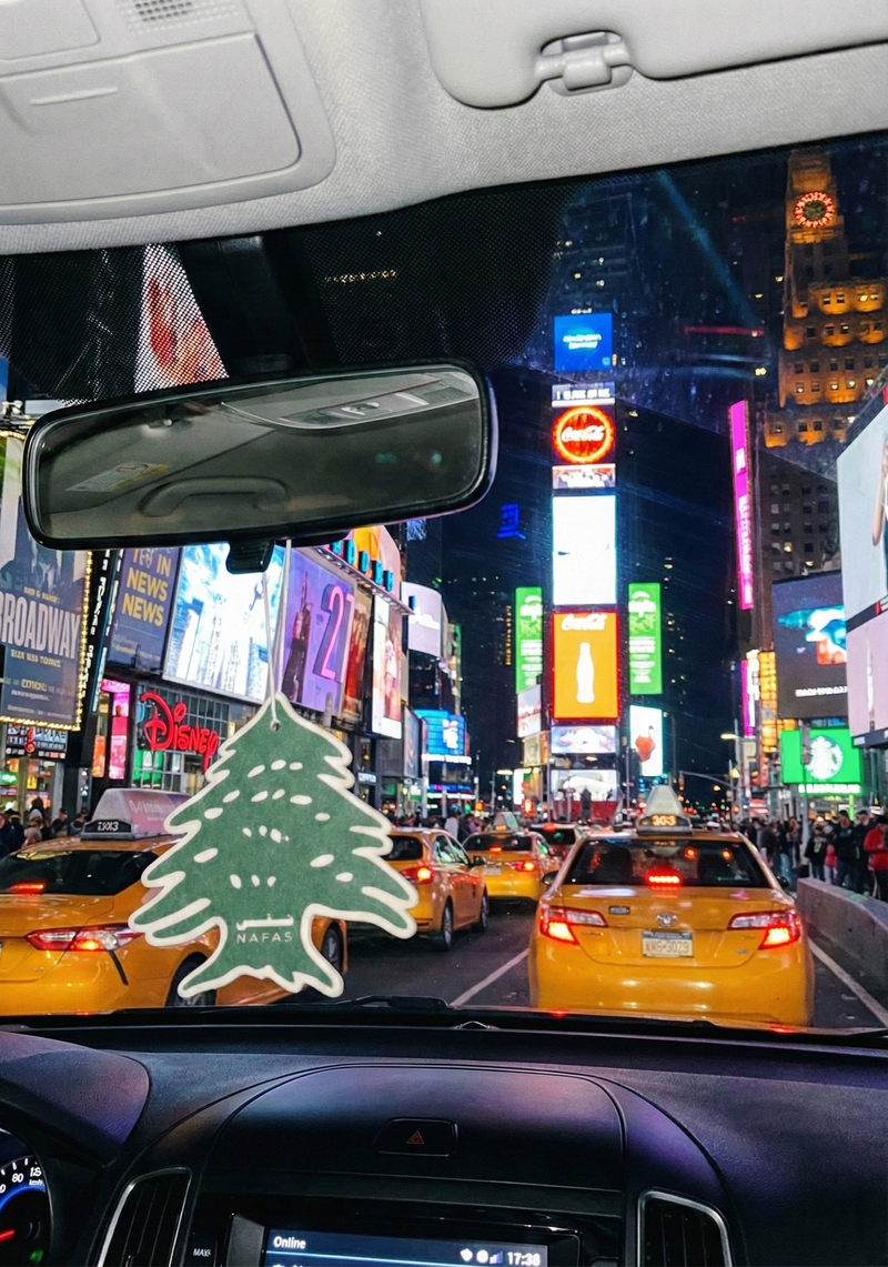 Times Square in New York City at night seen from inside a car with a Nafas Lebanese cedar air freshener hanging from the rearview mirror