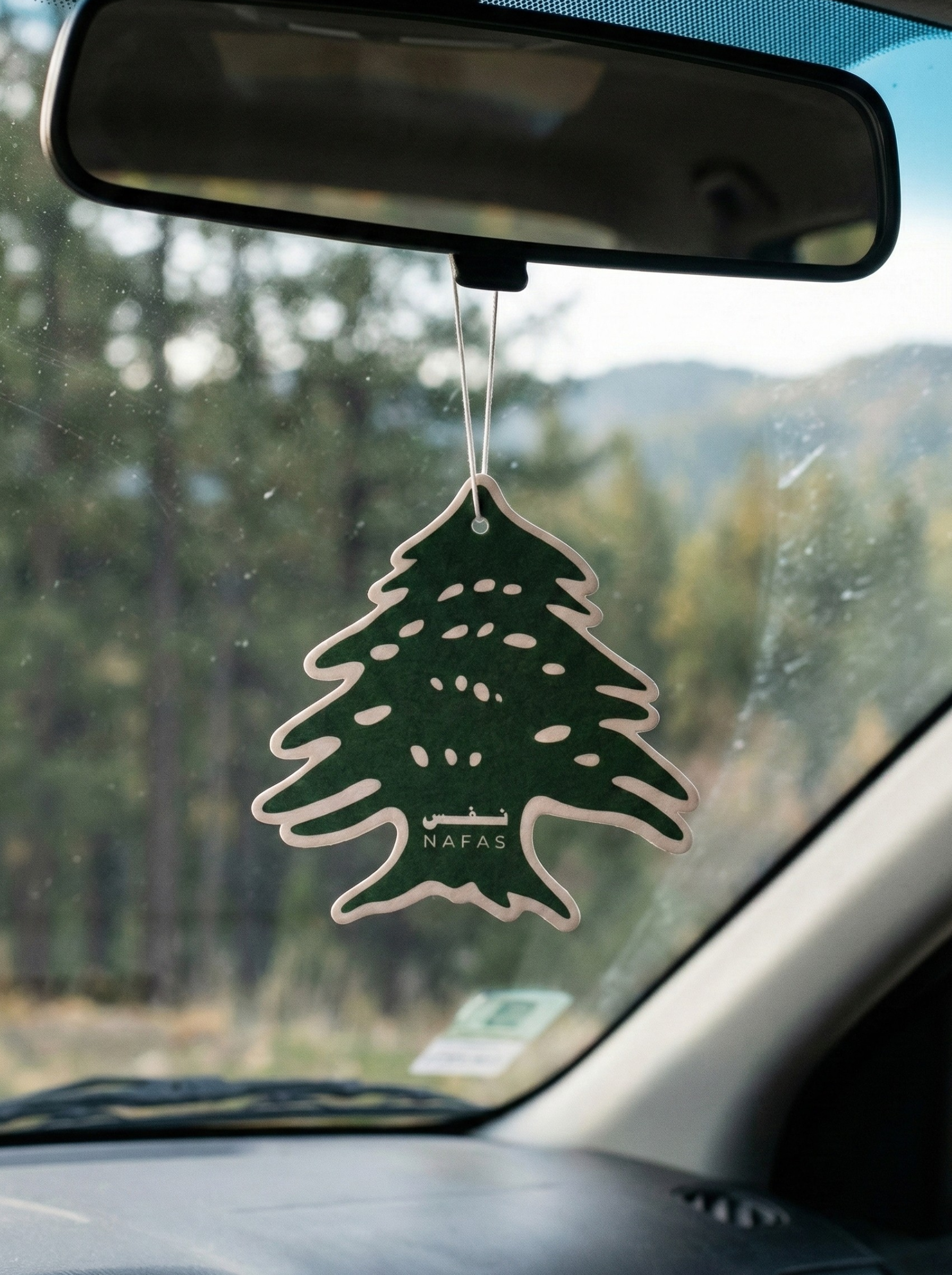 Nafas Lebanese cedar car air freshener hanging from a rearview mirror, Nafas dark green cedar-shaped Nafas freshener with forest view outside the windshield.